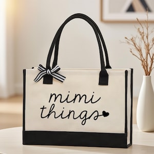 以下が含まれることがあります： 黒いトリムとハンドルが付いたクリーム色のトートバッグ。「mimi things」という言葉が筆記体フォントで書かれており、小さなハートのシンボルが付いています。白と黒のストライプのリボンが上部に付いています。
