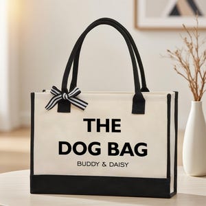 以下が含まれることがあります： 黒いトリムとハンドルが付いた、クリーム色のトートバッグ。「THE DOG BAG」の文字が大きく太字の黒で印刷され、その下に「BUDDY & DAISY」と書かれています。白と黒のストライプのリボンがハンドルに取り付けられています。