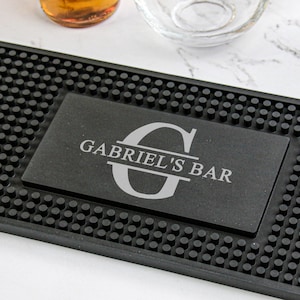 May include: A black rubber bar mat with a rectangular, personalized plaque that reads "GABRIEL'S BAR" in white lettering. A clear glass mug and a bottle of yellow liquid are visible in the background. The mat has a textured surface.