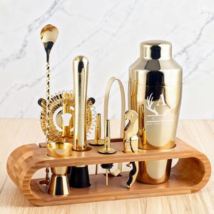 May include: A gold-colored bar tool set with a cocktail shaker, jigger, strainer, and other tools. The set is displayed on a bamboo wood stand. The shaker has an engraved initial and the words "ATHENA" and "ANY ROLE".