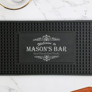 May include: Black rubber bar mat with a textured surface and a rectangular inset. The inset features the text "Welcome to MASON'S BAR Good Friends Good Drinks" in white, with decorative flourishes. The mat is designed for bar use.