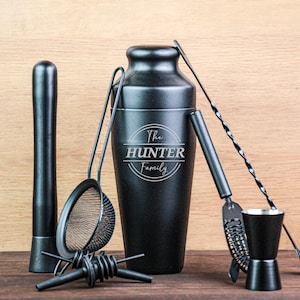 May include: A black cocktail shaker set including a muddler, strainer, jigger, bar spoon, and bottle opener. The shaker features the text "The Hunter Family". The set is presented on a wooden surface.