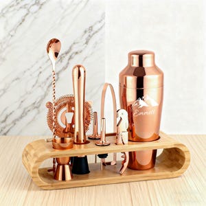 May include: A copper-colored bar tool set includes a cocktail shaker with the name "Emmett", a spoon, a strainer, a muddler, a jigger, a bottle opener, and other tools. The tools are displayed on a bamboo wood stand.