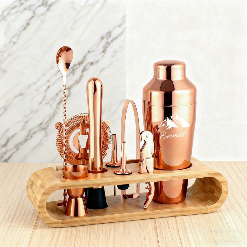 May include: A copper-colored bar tool set includes a cocktail shaker with the name "Emmett", a spoon, a strainer, a muddler, a jigger, a bottle opener, and other tools. The tools are displayed on a bamboo wood stand.