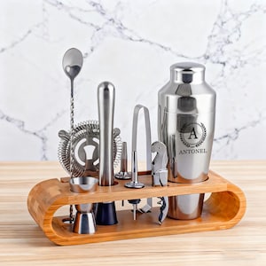 May include: A stainless steel bar tool set with a wooden holder. The set includes a cocktail shaker with the letter "A" and the name "ANTONEL", a strainer, a spoon, a jigger, a muddler, a bottle opener, and ice tongs.