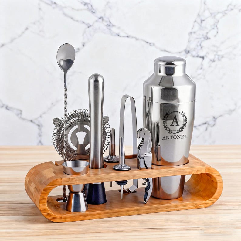 May include: A stainless steel bar tool set with a wooden holder. The set includes a cocktail shaker with the letter "A" and the name "ANTONEL", a strainer, a spoon, a jigger, a muddler, a bottle opener, and ice tongs.