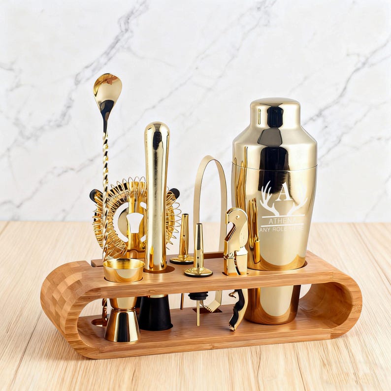 May include: A gold-colored bar tool set with a cocktail shaker, jigger, strainer, and other tools. The set is displayed on a bamboo wood stand. The shaker has an engraved initial and the words "ATHENA" and "ANY ROLE".