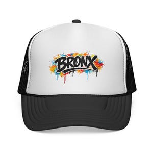 Puede incluir: Gorra de camionero en blanco y negro con un panel frontal blanco. La parte delantera presenta la palabra "BRONX" en negro, con un estilo graffiti estilizado, sobre un diseño de salpicaduras de pintura coloridas. La visera es negra.