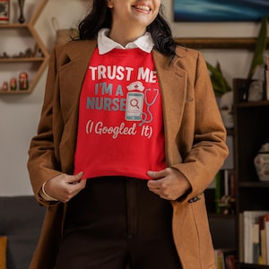 May include: A red sweatshirt with the text "TRUST ME I'M A NURSE (I Googled It)" in white and light blue. The sweatshirt features a stethoscope and a phone graphic. The person is wearing a brown blazer and dark brown pants.
