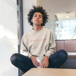 Puede incluir: Una sudadera beige con el texto "Choose Simplicity" en cursiva. La sudadera se combina con vaqueros azul oscuro. La persona sostiene una taza blanca con una pajita.