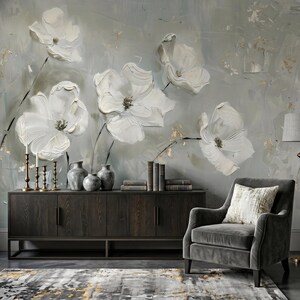 May include: A wall mural featuring large, textured white flowers against a soft gray background with gold accents. A dark wood sideboard holds three metallic candlesticks and two gray vases. A gray armchair with a patterned pillow sits nearby.