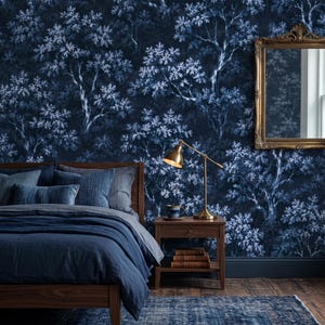 Op de afbeelding: Een slaapkamer met donkerblauw behang met een wit bomenpatroon, een houten bedframe met blauw beddengoed, een houten nachtkastje met een messing bureaulamp, een keramische mok, drie gestapelde boeken en een goudkleurige spiegel aan de muur.