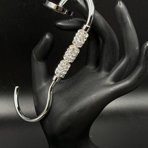 May include: A silver-toned metal purse hook featuring a curved design with three cylindrical beads encrusted with clear, sparkling rhinestones. The hook has a flat, circular base at the top end and a smooth, polished finish throughout the metal frame.