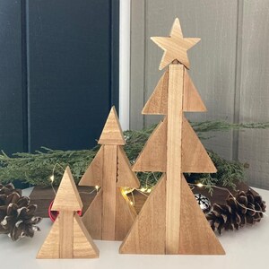 Può includere: Un set di quattro alberi di Natale in legno di varie dimensioni. Gli alberi sono costruiti in legno chiaro e hanno un design geometrico semplice. L'albero più grande ha una stella in cima. Gli alberi sono disposti su una superficie bianca con pigne e verde.