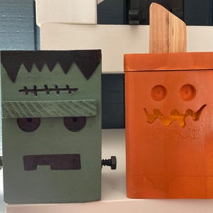 May include: Two wooden Halloween decorations: a green Frankenstein's monster and an orange jack-o'-lantern. The Frankenstein has black hair, stitched details, and bolt accents. The pumpkin has carved eyes and a mouth, with a wooden stem.