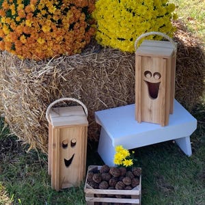 May include: Two wooden decorative lanterns with smiling face cutouts and rope handles. One lantern sits on a white bench, the other on the grass. A wooden crate filled with nuts and yellow flowers are in front of the bench. Orange and yellow mums and a bale of hay are in the background.