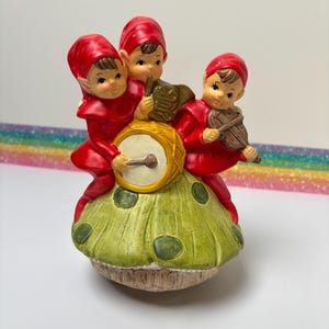 May include: A ceramic figurine of three musical elves in red outfits and hats. They are playing a drum, a horn, and a violin while standing on a green mushroom with brown spots. The figurine has a vintage aesthetic.