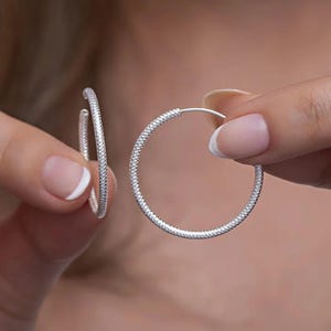 May include: A pair of silver hoop earrings. The earrings are round and feature a textured, studded design. One earring is held up by a person's hand, while the other is held up by another hand. The earrings are a classic jewelry item.
