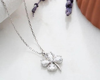 Puede incluir: Un collar de plata con un colgante de trébol de cuatro hojas, adornado con piedras preciosas transparentes. La cadena es delicada y el colgante está centrado. Ramas de lavanda y una maceta de terracota en el fondo, sobre una superficie blanca.