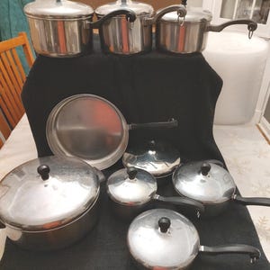 May include: A collection of stainless steel cookware, including pots, pans, and lids. The set features various sizes of pots with black handles and lids with black knobs. The cookware is arranged on a black surface, showcasing a complete kitchen set.