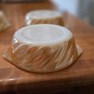 May include: Close-up of a set of vintage, iridescent glass bowls. The bowls have a swirl design on the sides and a smooth, flat top. The color is a warm, creamy peach. The bowls are sitting on a wooden surface.