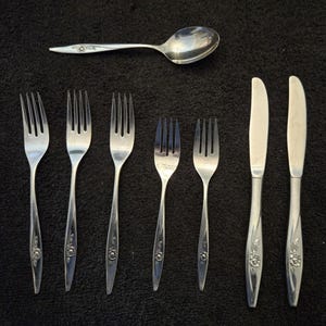 May include: A collection of silver-colored flatware, including forks, knives, and a spoon. The handles of the utensils feature a floral design. The spoon is positioned above the forks, with the knives to the right.