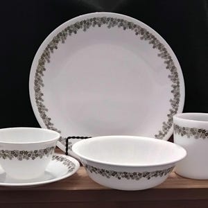 Op de afbeelding: Een wit servies bestaande uit een groot bord, een kom, een theekopje met schotel en een mok. Elk stuk is versierd met een delicaat bloemmotief in grijstinten en bruin langs de rand, wat een samenhangende en elegante uitstraling creëert.