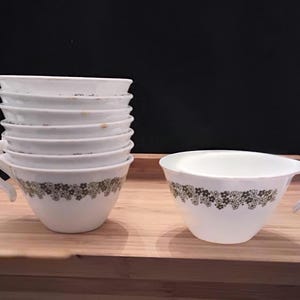 May include: A set of white ceramic teacups with a floral pattern in shades of green and brown. The cups have a curved handle and a wide rim. One cup is separate, and the others are stacked. The cups are on a wooden surface.