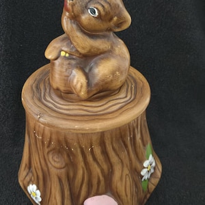 May include: A brown ceramic trinket box shaped like a tree stump, with a squirrel figurine on top. The box is decorated with white flowers and pink mushrooms. The squirrel has a red mouth and is holding its paw up to its face.