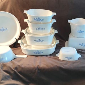 May include: A collection of white glass cookware featuring a blue floral pattern. The set includes a round plate, a handled saucepan, a stack of four rectangular baking dishes with glass lids, a small square dish, a bowl, and a loaf pan.