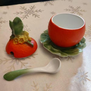 May include: A ceramic egg cup set with a tomato design. The set includes a red egg cup, a green leaf-shaped saucer, a matching lid, and a small spoon. The egg cup and lid have a vibrant orange color, while the spoon has a green and white gradient.