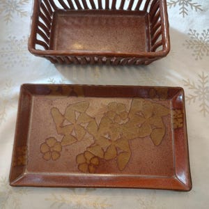 May include: Two-piece ceramic serving set. The top piece is a rectangular basket with vertical slats. The bottom piece is a rectangular tray with a floral design in shades of brown and tan. The set is on a white surface with snowflake patterns.