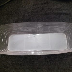 May include: A clear, rectangular plastic basket with a woven basket design on the sides. The basket has rounded corners and a smooth, flat bottom. The basket is empty and sits on a black surface.