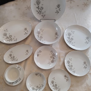 May include: A selection of white porcelain dinnerware featuring a grey rose design and gold edging. The set includes plates, bowls, and serving dishes, displayed on a patterned tablecloth. The dinnerware is likely vintage.