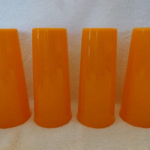 May include: Four tall, orange-colored drinking glasses. The glasses have a slightly flared base and a smooth, glossy finish. They are evenly spaced and arranged in a row, suggesting a set for serving beverages.