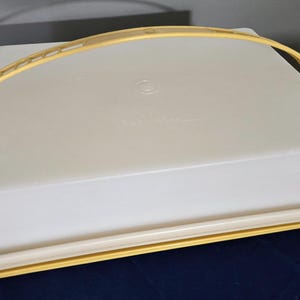 May include: A rectangular white plastic cake carrier with a yellow base and a matching yellow handle. The lid features a subtle embossed logo in the center. The carrier is designed for transporting baked goods and has a secure locking mechanism.