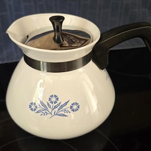 May include: A white teapot with a rounded body and a black handle. The teapot features a blue floral pattern on the front, a metallic silver band around the neck, and a black lid with a central knob. The design is simple and functional.