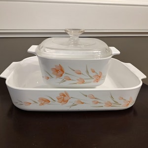 May include: A white rectangular baking dish and a smaller white square casserole dish with a clear glass lid. Both pieces feature a repeating pattern of orange tulip-like flowers with green stems and leaves printed along the exterior sides.