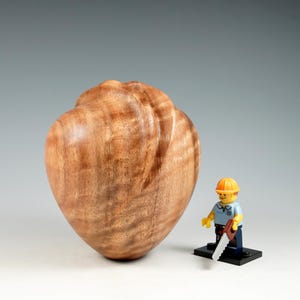 May include: A heart-shaped wooden sculpture, hand-carved with visible wood grain. The sculpture has a reddish-brown hue. A Lego figure with a saw is positioned next to it for scale. This is likely a decorative item, possibly for the home.