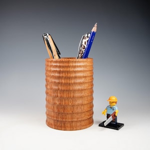 May include: A cylindrical wooden pencil holder with a ribbed, textured exterior surface. Two pens are inserted into the top. A small plastic construction worker figurine wearing a yellow hard hat and holding a saw stands next to the holder.