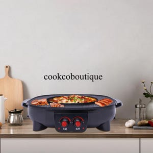 Könnte beinhalten: Ein schwarzer Elektrogrill mit runder Kochfläche, mit Pizza und gegrilltem Fleisch. Der Grill hat zwei rote Bedienknöpfe und den Text "cookcoboutique" an der Wand dahinter. Der Grill steht auf einer Holztheke.