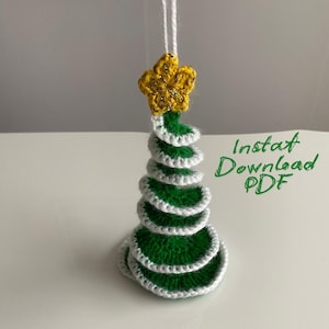 May include: Crocheted Christmas tree ornament. The tree is green with white trim and topped with a gold star. The text "Instat Download PDF" is written in green on the right side of the image. The ornament hangs from a white string.