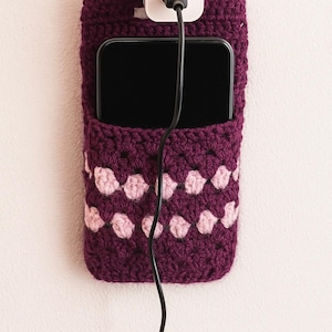 May include: A handmade, crochet phone holder in a deep purple hue, featuring a decorative pattern of pink stitches. The holder is mounted on a wall and has a phone inside, with a black charging cable plugged into a white adapter.