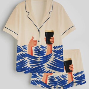 May include: Cream-colored pajama set with a short-sleeved button-up shirt and matching shorts. The set features a blue wave design and illustrations of hands holding a pint of dark beer. The shirt and shorts are trimmed with navy blue piping.
