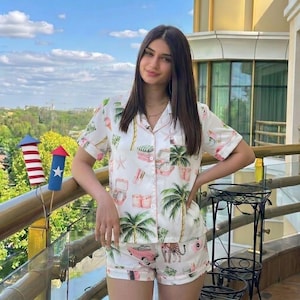 May include: White pajama set with a tropical print featuring palm trees, pink cars, and a leopard. The short-sleeved top and shorts have pink trim. The set is made of a lightweight fabric.