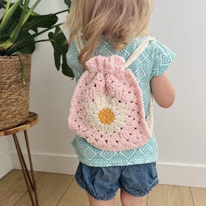 May include: A light pink crochet drawstring backpack featuring a large white daisy motif with a yellow center. The bag is constructed with a granny square pattern and includes thick, cream-colored braided cord straps for carrying on the shoulders.