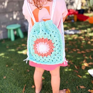 May include: A crochet drawstring backpack featuring a light blue granny square pattern with a central flower motif in shades of white, light pink, and coral. The bag includes thick white crocheted straps that function as a closure for the main compartment.