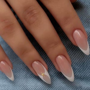 Op de afbeelding: Amandelvormige nagels met een klassieke French manicure, voorzien van een transparante nude basis en strakke witte randen. Een kleine, goudkleurige sterbedel is in het midden van de nagel van de ringvinger geplaatst voor een accent.
