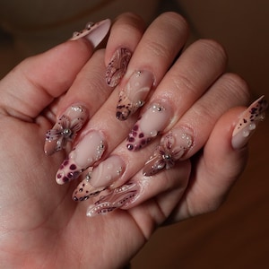 May include: Long almond-shaped nails featuring a nude base with intricate brown and white hand-painted floral patterns, swirling lines, and spotted designs. The nail art is embellished with small silver metallic beads and clear raised 3D accents.