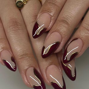 Può includere: Primo piano di unghie curate con una combinazione di bordeaux e nude, accentuata da linee dorate. Le unghie a mandorla presentano un design a french manicure con un tocco moderno. È visibile anche un anello d'oro con pietre trasparenti.